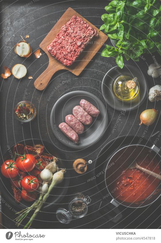 Ingredients for sauce Bolognese: minced meat, basil, tomatoes, garlic, onion, oil, kitchen utensils and spices on dark kitchen table. Cooking italian food at home. Top view.