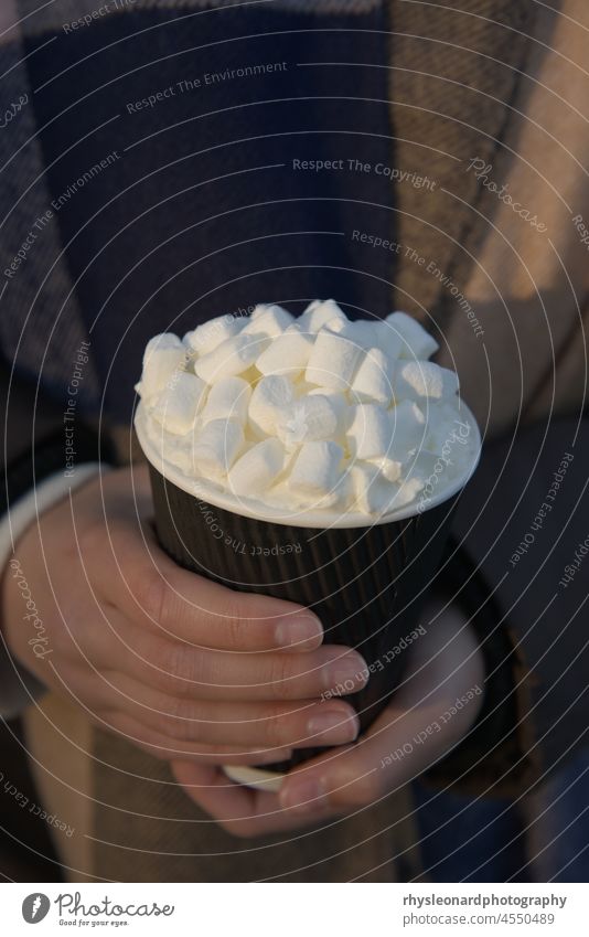 Hot take away beverage with an abundance of marshmallows and cream, held in the hands of a young woman. Background of woollen winter scarf texture. Warm evening sunlight lights the scene from the side.