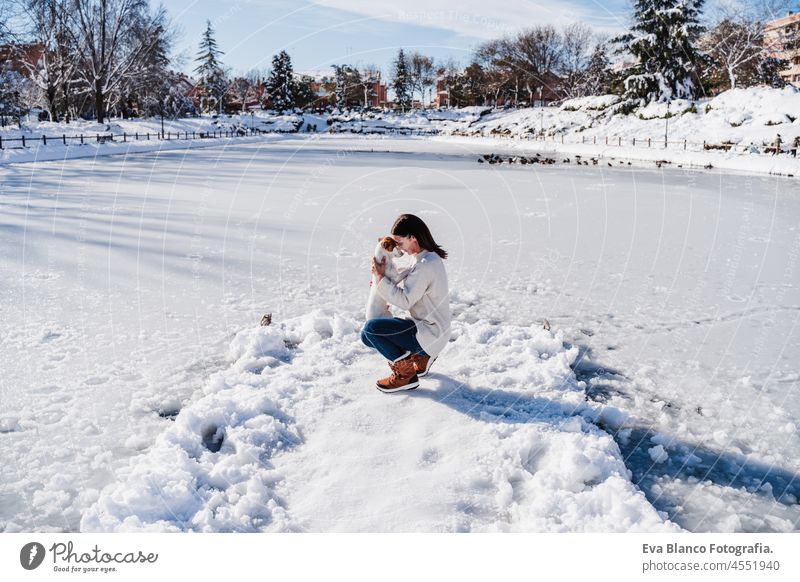 caucasian woman hugging cute jack russell dog while standing on snowy pier by frozen lake during winter on sunny day. winter landscape in city park dock cold