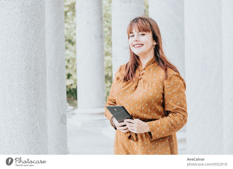 Portrait of a young red head woman looking straight to camera while smiling. Windy day. Autumnal dress colorful days. Day at the park, connect wireless video call. Copy space