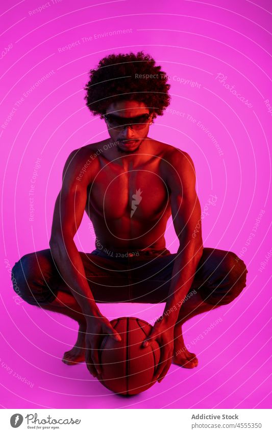 Black shirtless man sitting with basketball naked torso muscular sportsman appearance player neon squat male african american black curly hair sunglasses young