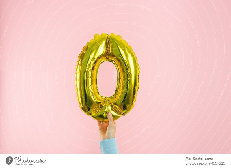 Woman's hand in blue jumper holding golden globe in the shape of number 0 on pink background. Copy space. zero balloon copy copy space symbol woman bracelet