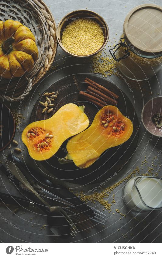 Butternut squash halves on plate with millet, cardamom, cinnamon, bowls and wooden basket on pale grey concrete kitchen table. Preparing healthy vegan food with seasonal ingredients at home. Top view