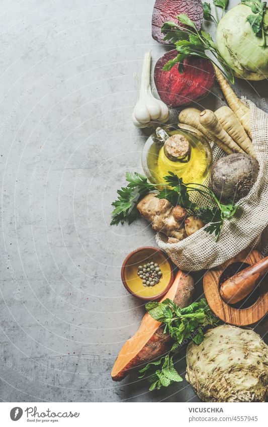 Food background with various root vegetables and ingredients: parsnip, sweet potato, kohlrabi, beetroot, celery, garlic and olive oil on grey table. Seasonal cooking at home. Top view. Border