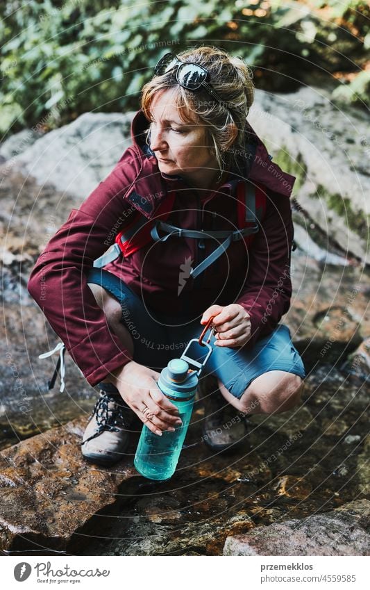 Woman taking pure water to bottle from mountain stream during trekking in mountains adventure trip travel hiking thirsty vacation journey crouching summer