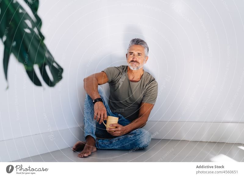 Pensive senior male sitting on floor with cup of tea man pensive drink think dream chill rest apartment wall pastime style loft beard handsome contemplate mug