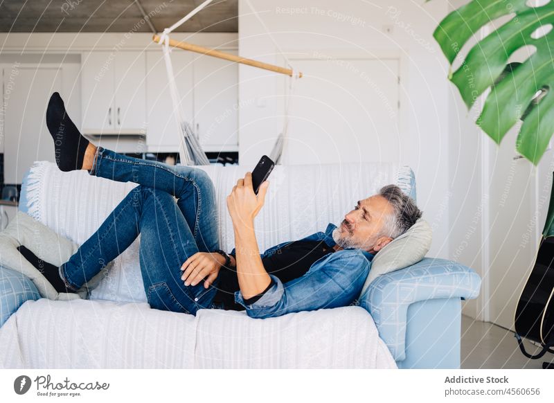 Senior man browsing cellphone lying down on couch smartphone mobile sofa using style loft chill rest pastime living room aged jeans modern free time denim relax