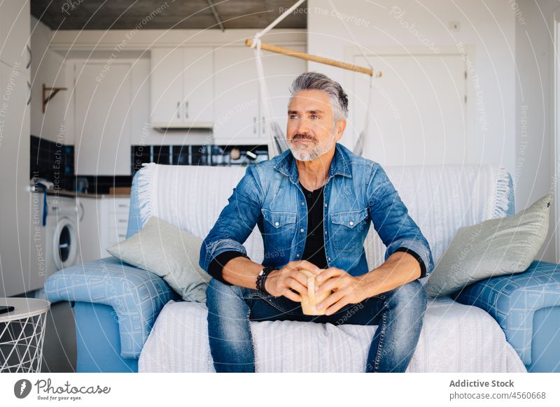 Aged wistful man with cup of tea sitting on sofa drink comfort calm couch loft thoughtful chill rest mug unshaven ponder pastime aged alone think beverage jeans