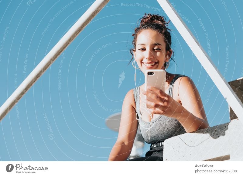 Young woman with curly hairstyle sitting on urban floor.Leggings clothing and top Fitness concept.Resting after workout fitness. Sportswear, smart phone, headphones.Active lifestyle, motivation power.