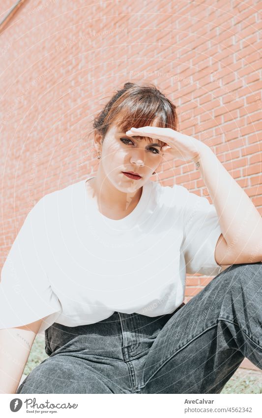 Young beautiful red head looking serious to camera hipster woman .Trendy girl in summer T-shirt and black jeans .Serious and pensive female posing in the street near a brick wall.