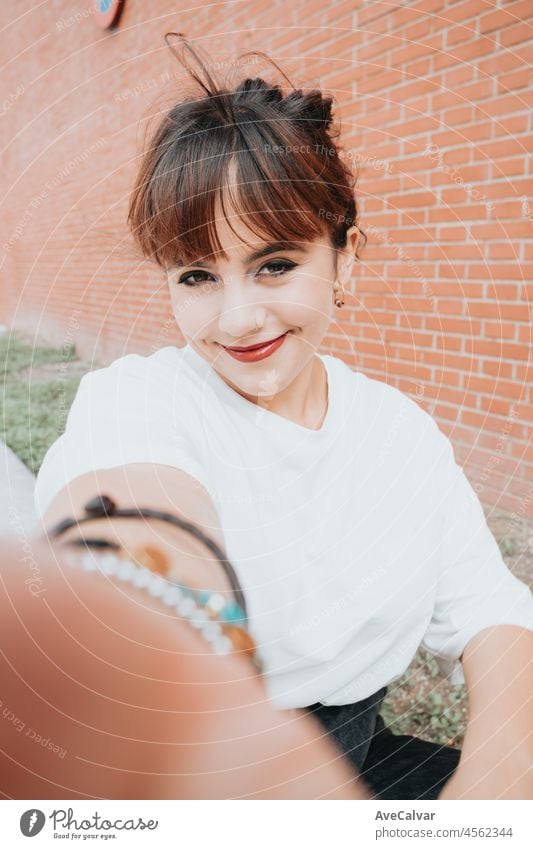 Young beautiful red head smiling hipster woman taking selfie .Trendy girl in summer T-shirt and black jeans .Positive female posing in the street near a brick wall.