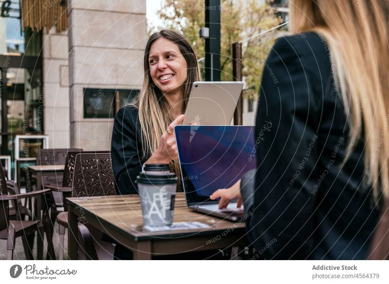 Cheerful businesswoman near faceless coworker at table with gadgets businesswomen cafe terrace tablet laptop online browsing modern street spend time city