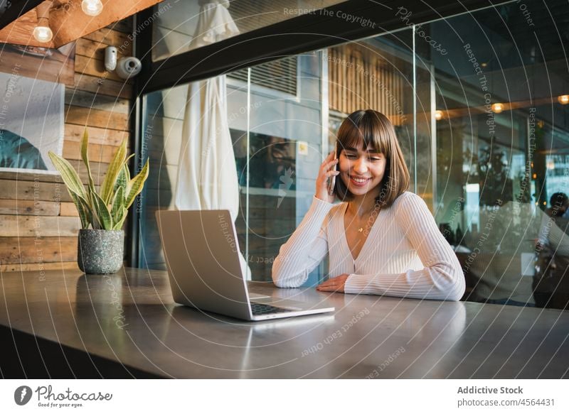 Smiling woman talking on smartphone in cafeteria with laptop freelance work mobile gadget device computer using netbook cellphone call speak communicate