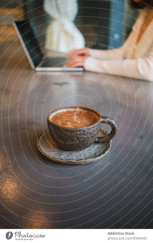 Cup of cappuccino placed on table near woman with laptop delicious coffee gadget cup drink typing tasty using freelance browsing cafe surfing fresh ceramic work