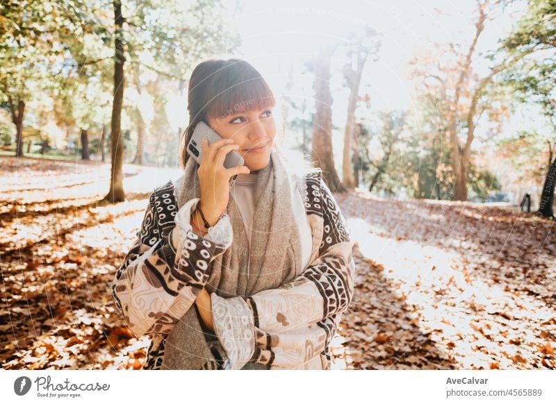 Happy girl using smart phone during Autumn in a park taking selfie, woman talking on mobile in fall. Video call outdoors, receiving good news. person female