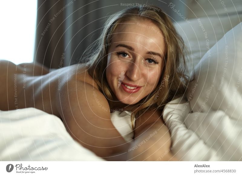 Nude portrait of a young, blond, long-haired woman lying crosswise in bed on her stomach and smiling into the camera Youthfulness salubriously Facial expression