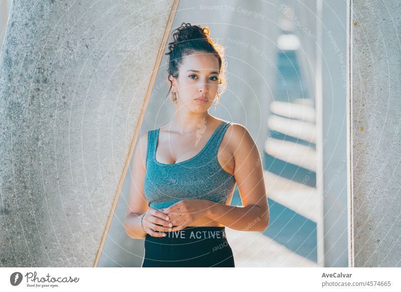 Serious portrait of a youn African american athletic woman, curly hairstyle,resting after running for a competition in the city. Sunset time with shadows. Serious woman wants to win the race