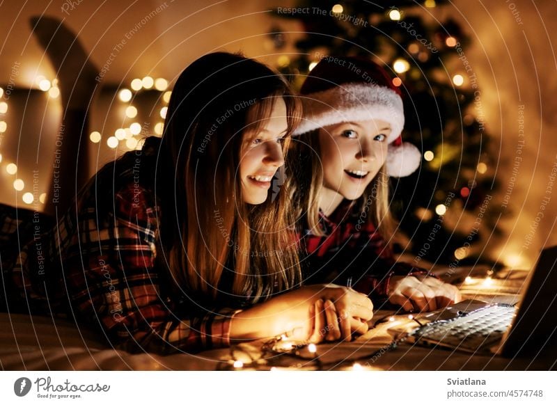 Two charming sisters lie on the bed with a laptop on Christmas or New Year's eve and congratulate their friends and relatives online. Christmas greetings online, virtual party for Christmas