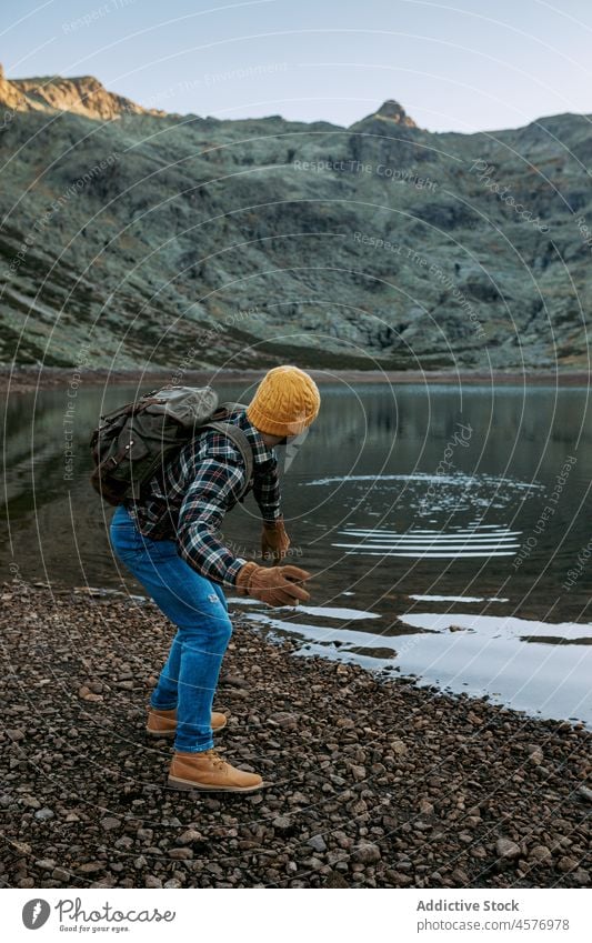 Unrecognizable traveler skimming stone on water surface man hiker mountain lake shore waterside throw nature journey ridge rough formation coast tourist