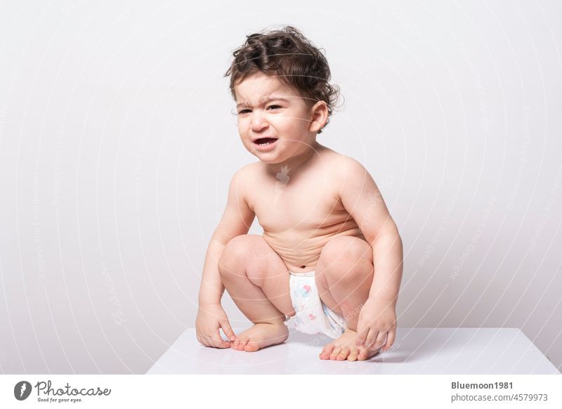 One year old baby wearing a diaper and squatting on a white table - crying expression on his face Caucasian adorable alone backdrop beautiful care one year old