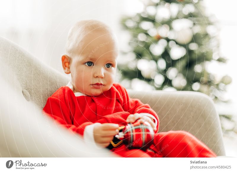 A child dressed as Santa Claus is sitting in a chair next to a Christmas tree on Christmas Eve christmas baby santa white claus christmas eve christmas time