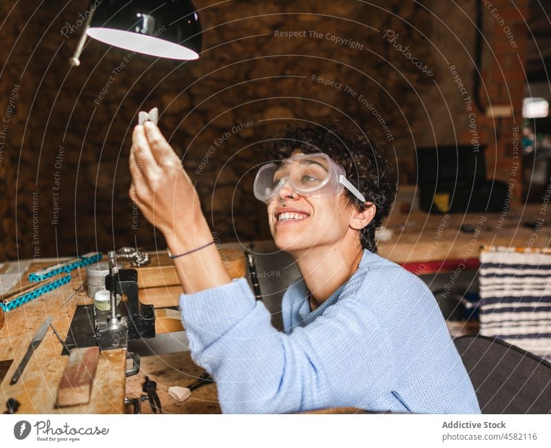 Happy goldsmith examining jewelry in hand craftswoman jeweler accessory lamp workshop precious smile examine workbench small business cheerful female