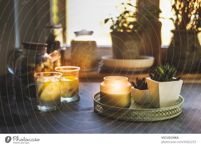 Cozy kitchen with lemon tea in glass cups, tea pot, candles and small potted plants at window background. Cold season with warm beverages. Front view. cozy