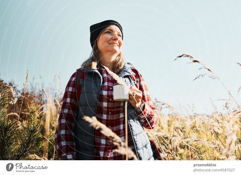 Smiling woman relaxing and enjoying the coffee during summer trip. Woman standing on trail and looking away holding cup of coffee. Woman with backpack hiking through tall grass along path in mountains