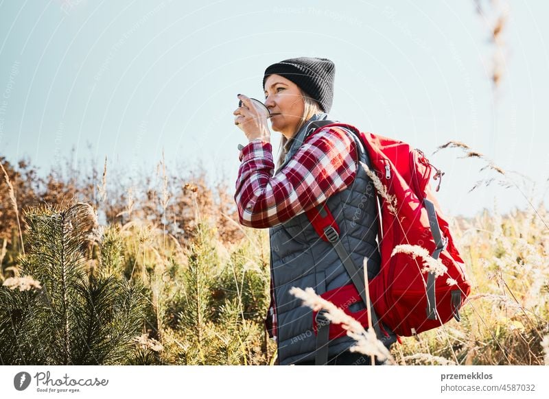 Woman taking break and relaxing with cup of coffee during summer trip. Woman standing on trail and looking away. Female with backpack hiking through tall grass along path in mountains