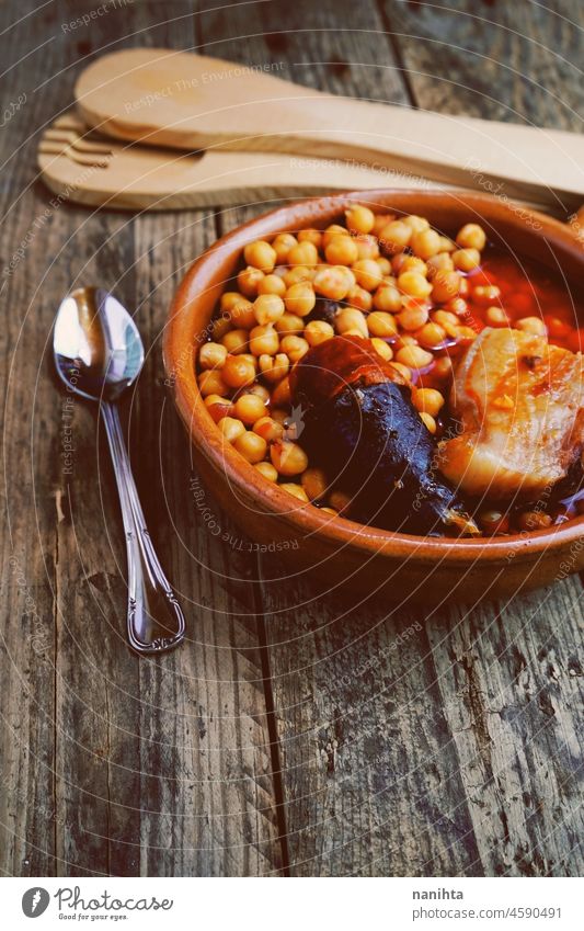 Chickpeas, sausage and bacon in a crockpot by wooden spoon and fork, and a little metal spoon. Typical food from Madrid, Spain, with a rustic wooden board as a background.