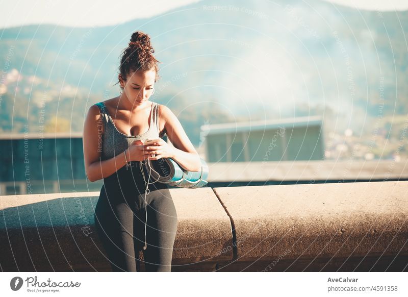 Smiling young african woman holding and using smartphone during workout while sitting outside holding yoga mat listening to music and chatting outdoors . New habits life style. Urban sport sunset