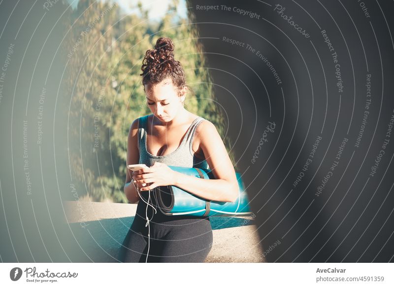 Smiling young african woman holding and using smartphone during workout while sitting outside holding yoga mat listening to music and chatting outdoors . New habits life style. Urban sport sunset