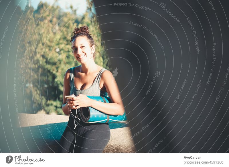 Smiling young african woman holding and using smartphone during workout while sitting outside holding yoga mat listening to music and chatting outdoors . New habits life style. Urban sport sunset