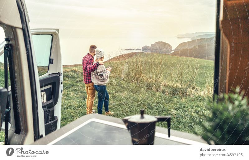 Unrecognizable couple traveling in camper van looking coastline landscape unrecognizable sea morning dawn coffee cup copy space sunlight romantic