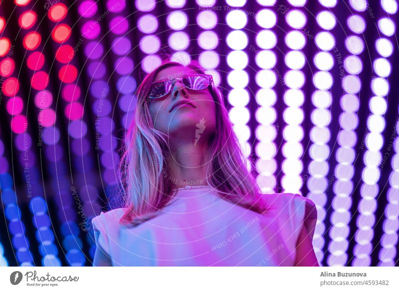 Teen hipster girl in stylish glasses standing on purple tunnel with neon light wall background, female teenager fashion model pretty young woman looking at night club city light glow