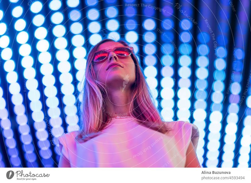 Teen hipster girl in stylish glasses standing on blue tunnel with neon light wall background, female teenager fashion model pretty young woman looking at night club city light glow