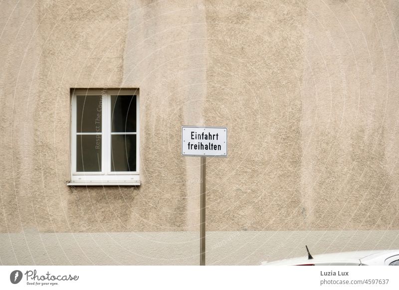 Sign 'Keep driveway clear' in front of a shabby house wall and in front of it the roof of a car whose driver ignored the notice keep the driveway clear