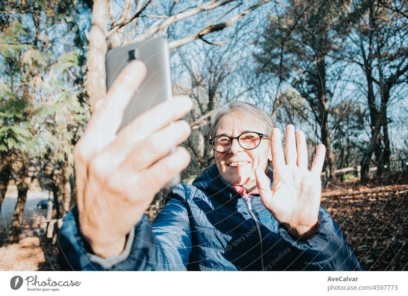 Happy mature woman having a video chat through smart phone while a day at the forest during a travel to treck. Copy space sitting on a wood bench. Active senior woman healthy life style. Calling grandchildren