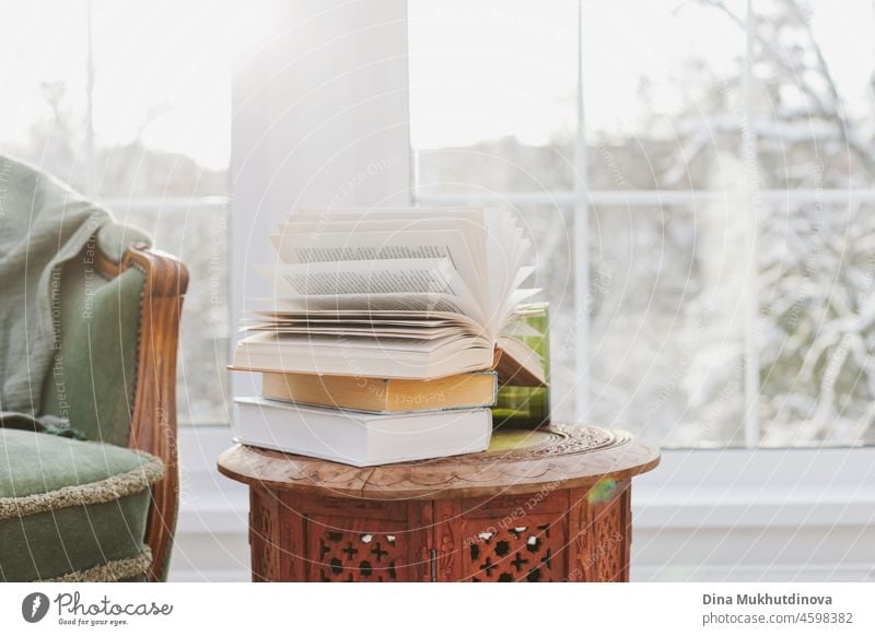 A stack of books on the wooden coffee table near armchair in the room with snowy trees landscape window view. Cozy home in winter. education literature read