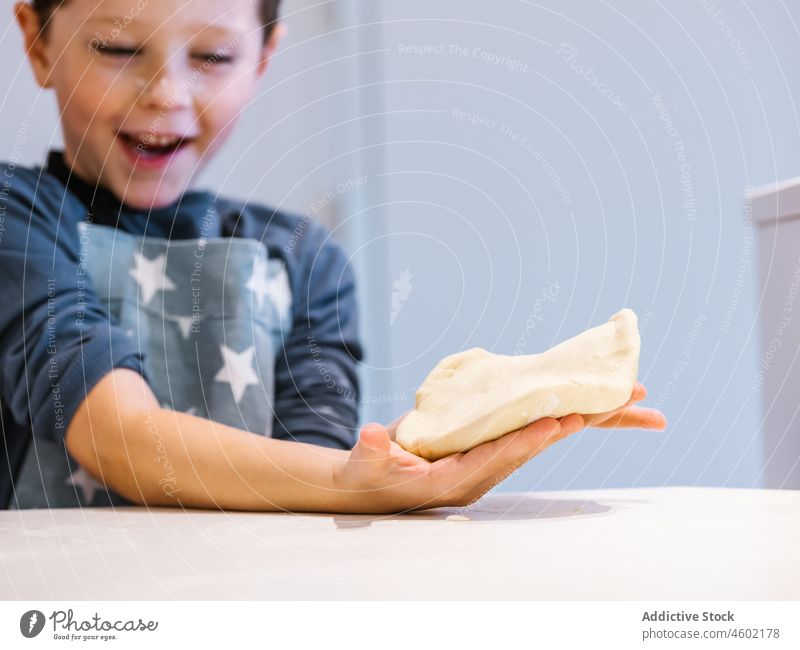 Cute boy in apron and with flour on face preparing dough for pastry while cooking in kitchen at home kid knead having fun helper tear process bake food domestic