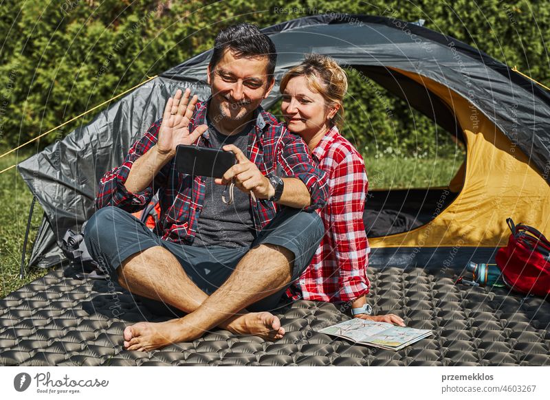 Couple having video call with friends using smartphone while sitting in tent at camping. People relaxing in tent during summer vacation trip adventure campsite