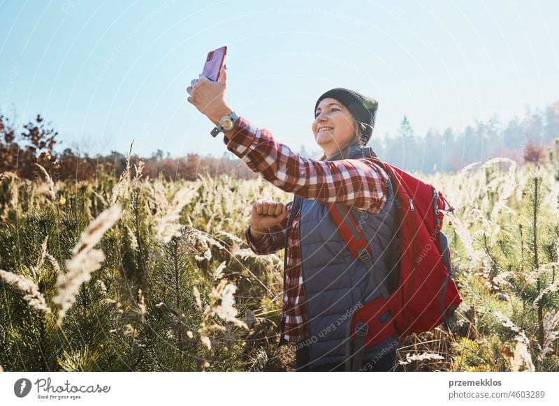 Young woman making video call sending greetings from vacation trip in mountains. Woman with backpack hiking through tall grass along path on meadow adventure