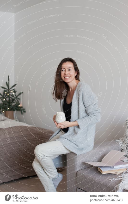 Millennial woman in cozy apartment holding a cup of coffee sitting on the couch near open books on holiday season lifestyle millennial drinking coffee relax