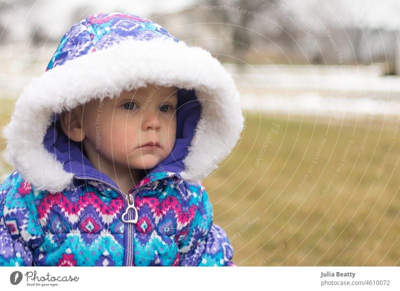 18 month old baby girl in a colorful patterned snow suit and hood looks away from the camera with a serious expression; waiting for the snow to fall snowsuit