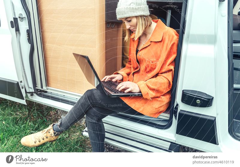 Woman teleworking sitting in the door of campervan woman telecommuting laptop trip remote work enjoying nature people cheerful looking around caravan wool cap