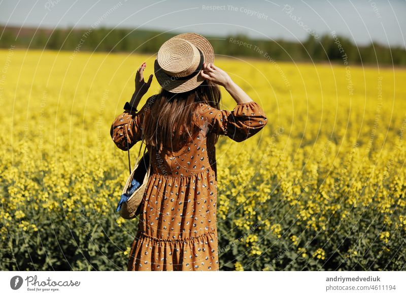 Rear view. beautiful young woman in a dress holding a hat and walking in a rapeseed field for summer, view from the back. copy space. summer holiday concept