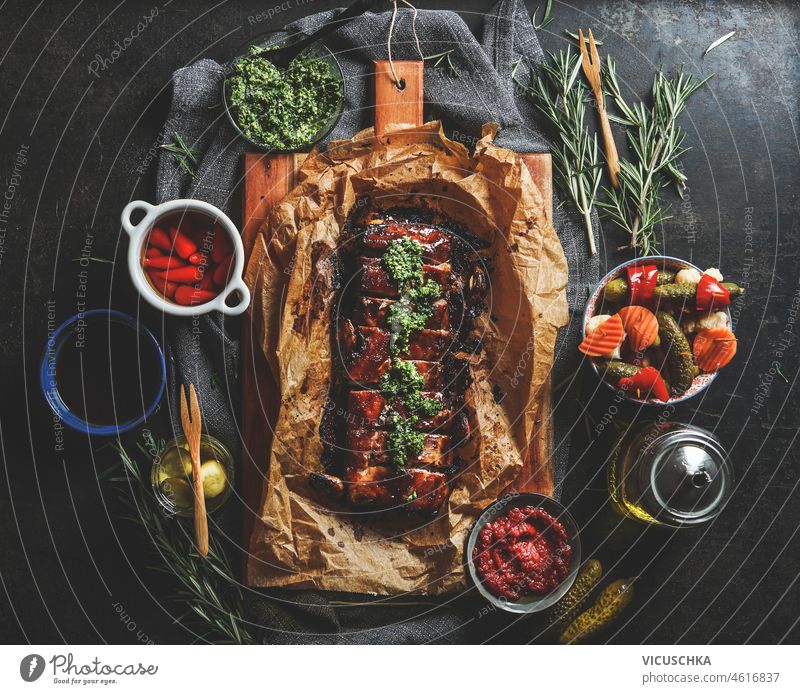 Roasted BBQ ribs with homemade pesto, pickled vegetables, sauces and rosemary on dark concrete kitchen table. Top view. roasted bbq flavorful meat dinner