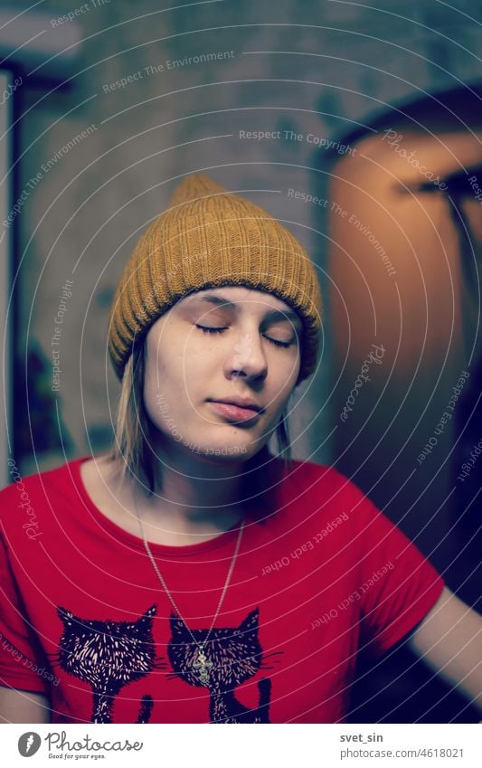 Portrait of a girl with her eyes closed in a mustard-colored knitted hat and a red T-shirt indoors with artificial lighting. young portrait Closed eyes person