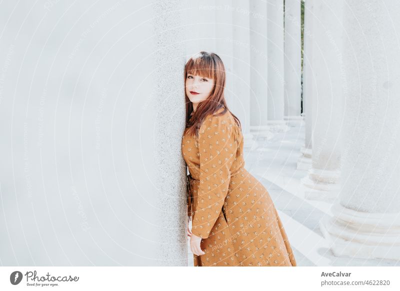 Young red head woman resting his face against a wall stressed and tired about the city life while looking serious to camera. Wearing trendy and modern clothes autumnal winter dress.