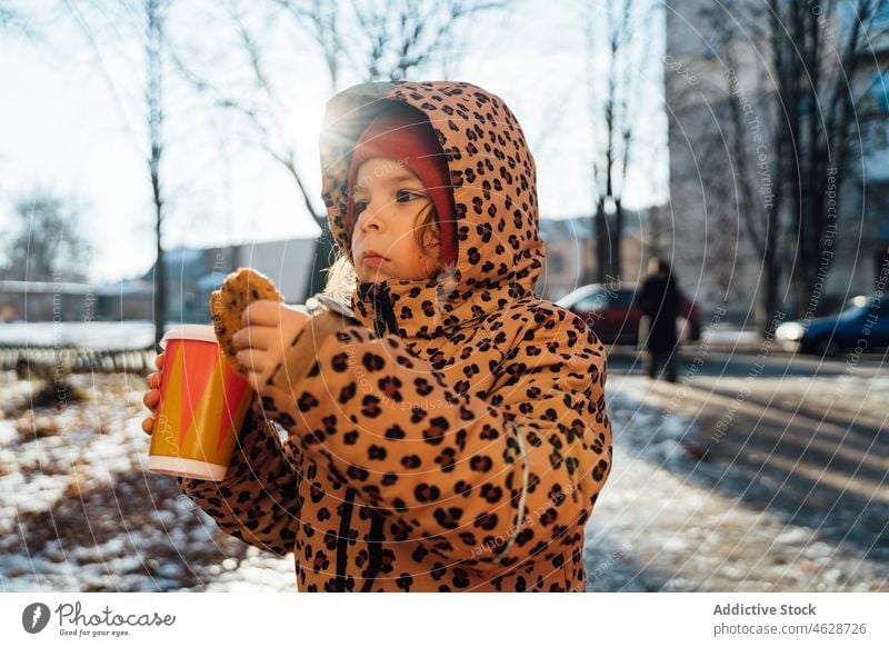 Cute girl with takeaway beverage on winter day kid cookie hot drink street city cold childhood dessert outerwear season snow coffee warm clothes serious